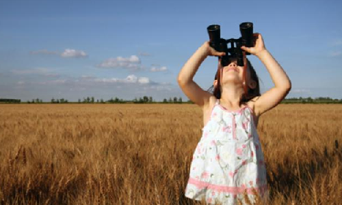 Image of a tyoung female child in a summer dress, standing in a grassy field looking into the distance with her binoculars.