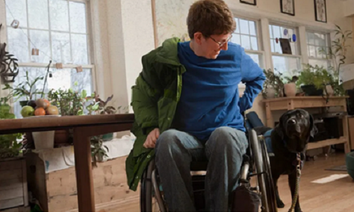 A woman preparing to leave her house. She wears warm clothing and is seated in a wheelchair. She smiles at her friemndly pet dog.